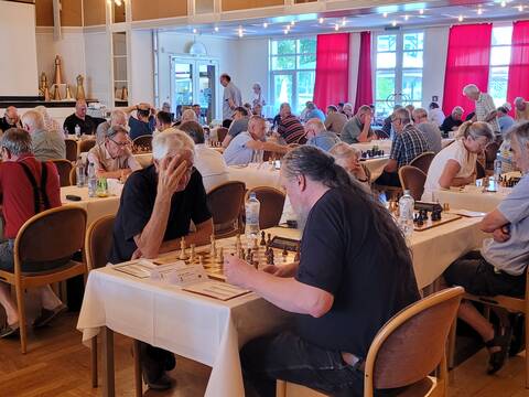 Auf dem Bild sind zahlreiche Schachspieler zu sehen, die sich an Tischen gegenüber sitzen und Schachpartien spielen.