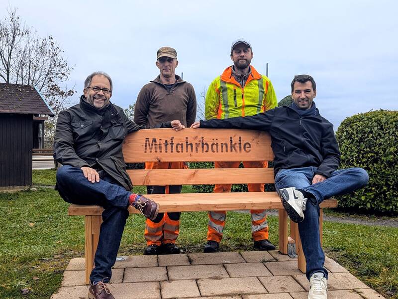 In der Bildmitte ist eine Holzbank mit dem in die Lehne eingebrannten Text Mitfahrbänkle zu sehen. Links außen auf der Bank sitzt ein Mann, ebenso rechts außen. In der Bildmitte hinter der Lehne stehen zwei Männer, die aufgrund ihrer Kleidung als Bauhof-Mitarbeiter zu erkennen sind. Bank