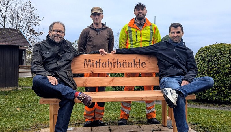 In der Bildmitte ist eine Holzbank mit dem in die Lehne eingebrannten Text Mitfahrbänkle zu sehen. Links außen auf der Bank sitzt ein Mann, ebenso rechts außen. In der Bildmitte hinter der Lehne stehen zwei Männer, die aufgrund ihrer Kleidung als Bauhof-Mitarbeiter zu erkennen sind. Bank