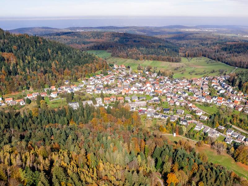 Das Bild zeigt ein Dorf, umgeben von dichtem, herbstlich gefärbtem Wald und sanften Hügeln.