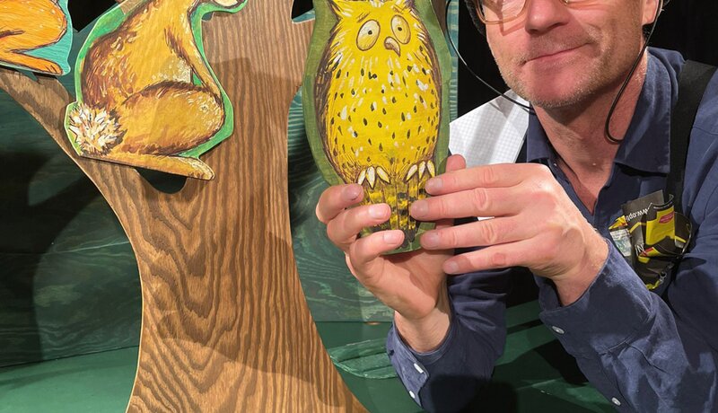 Ein Mann mit Brille und Mütze hält eine Eulenfigur aus Holz neben einem Baum mit einem Eichhörnchen und einem Hasen aus Pappe. | © marotte-Theater Karlsruhe