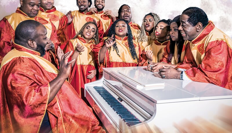 Eine Gruppe von Personen in roten und goldenen Roben singt enthusiastisch um ein weißes Klavier herum. | © Golden Voices of Gospel