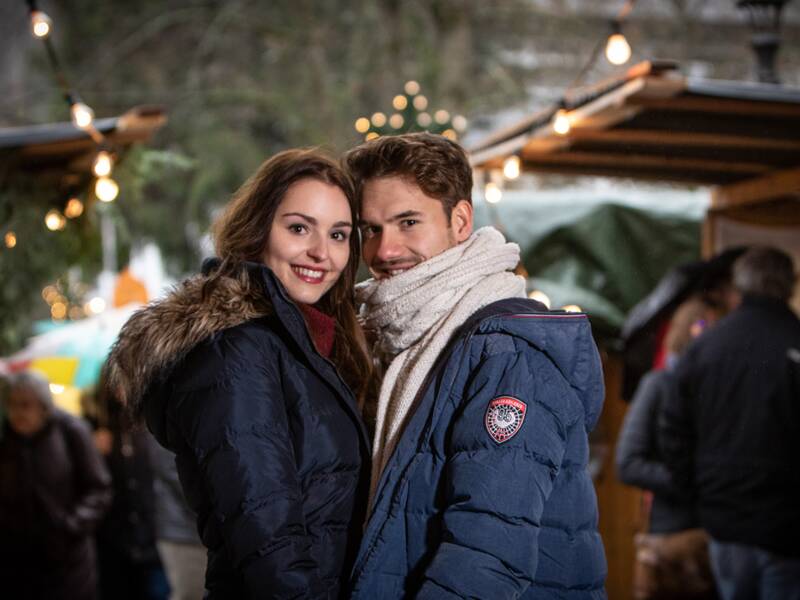 Ein junges Paar beim Besuch eines Adventsmarktes. Sie stehen dicht beiander, halten sich an den Händen und schauen in die Kamera. Der Adventsmarkt im Hintergrund ist verschwommen zu erkennen.