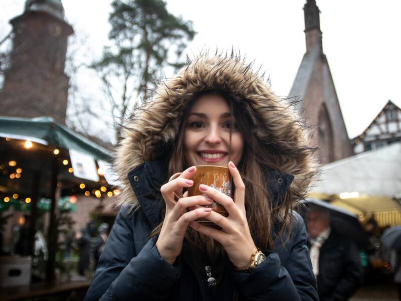 Eine junge Frau hält einen Glühweinbecher an ihr Gesicht und lächelt in die Kamera. Sie steht inmitten eines Adventsmarktes, dessen Lichter und Stände im Hintergrund unscharf zu erkennen sind.