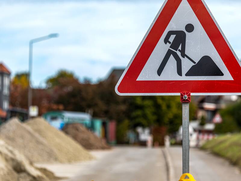 Ein Verkehrsschild, das auf eine Baustelle hinweist. Im Hintergrund befinden sich Sandhäufen auf einer Straße.