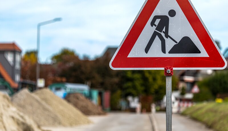 Ein Verkehrsschild, das auf eine Baustelle hinweist. Im Hintergrund befinden sich Sandhäufen auf einer Straße.