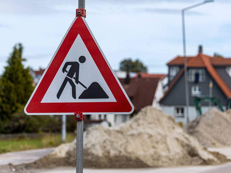 Ein Verkehrsschild, das auf eine Baustelle hinweist. Im Hintergrund befinden sich Sandhäufen auf einer Straße.