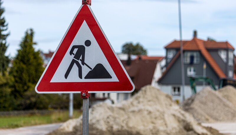 Ein Verkehrsschild, das auf eine Baustelle hinweist. Im Hintergrund befinden sich Sandhäufen auf einer Straße.