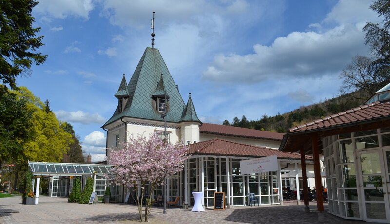 Zu sehen ist links im Bild ein von außen Kurhaus und rechts angeschnitten im Vordergrund ein verglaster Trinkpavillon.