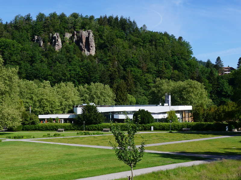 Ein modernes Gebäude mit flachem Dach steht vor einem bewaldeten Hügel mit Felsformationen, umgeben von einer grünen Wiese und Gehwegen.