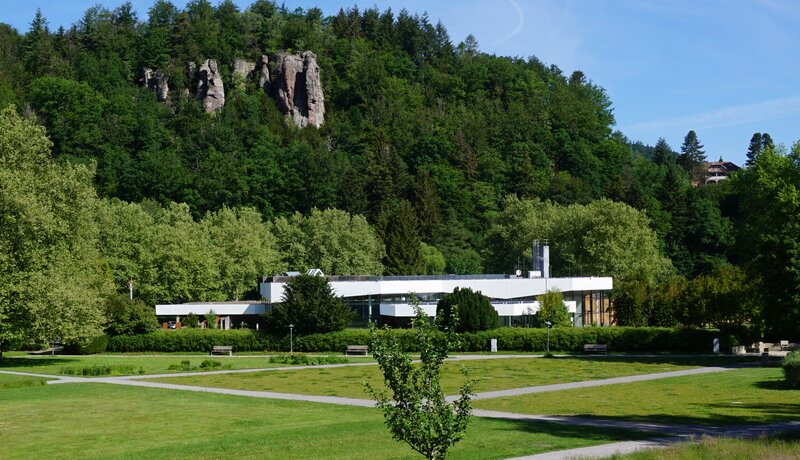 Ein modernes Gebäude mit flachem Dach steht vor einem bewaldeten Hügel mit Felsformationen, umgeben von einer grünen Wiese und Gehwegen.