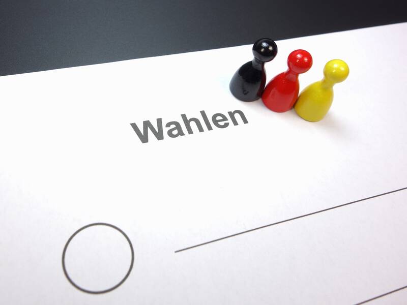 Ein Blatt Papier mit der Aufschrift "Wahlen" darauf, daneben stehen drei Spielfiguren in den Farben Schwarz, Rot und Gelb.