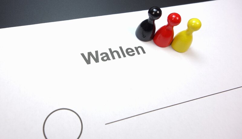 Ein Blatt Papier mit der Aufschrift "Wahlen" darauf, daneben stehen drei Spielfiguren in den Farben Schwarz, Rot und Gelb.