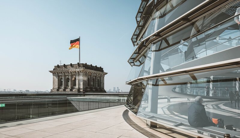 Ein Mann sitzt in der gläsernen Kuppel des Reichstagsgebäudes in Berlin, während im Hintergrund die deutsche Flagge flattert.